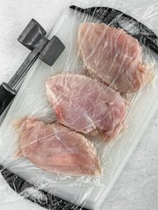 A cutting board is lined with plastic wrap then layered with chicken breasts then more plastic wrap overtop the chicken. A meat tenderizer sits besides the cutting board to tenderize the meat.