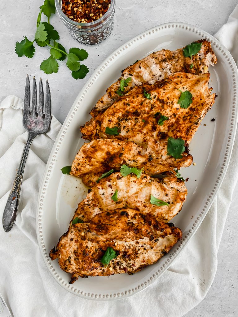 A platter is filled with the best grilled chicken breasts. Green parsley garnishes the chicken with more sitting to the side. A large serving forks sit beside the platter. There's a white linen beneath the platter and a jar of red pepper flakes to the side of it.