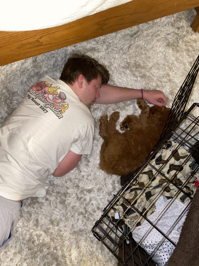 boy laying on white fluffy rug next to dog crate while puppy sleeps in his arms