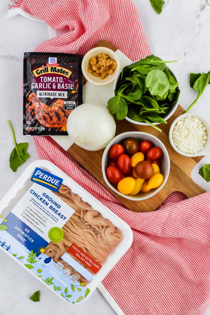 ingredients to make chicken burgers including spinach, garlic, feta, white onion, seasoning, and ground chicken on cutting board and red dish towel with white background