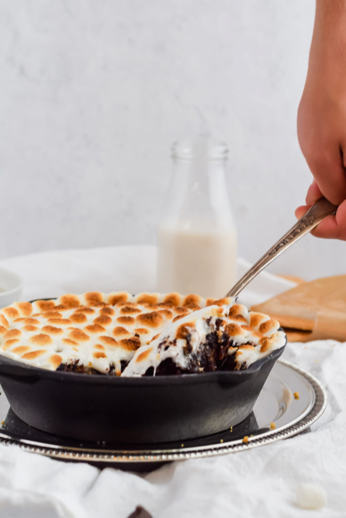 side shot of s'mores brownie skillet in cast iron skillet on silver tray with a spoon taking out a serving on white background