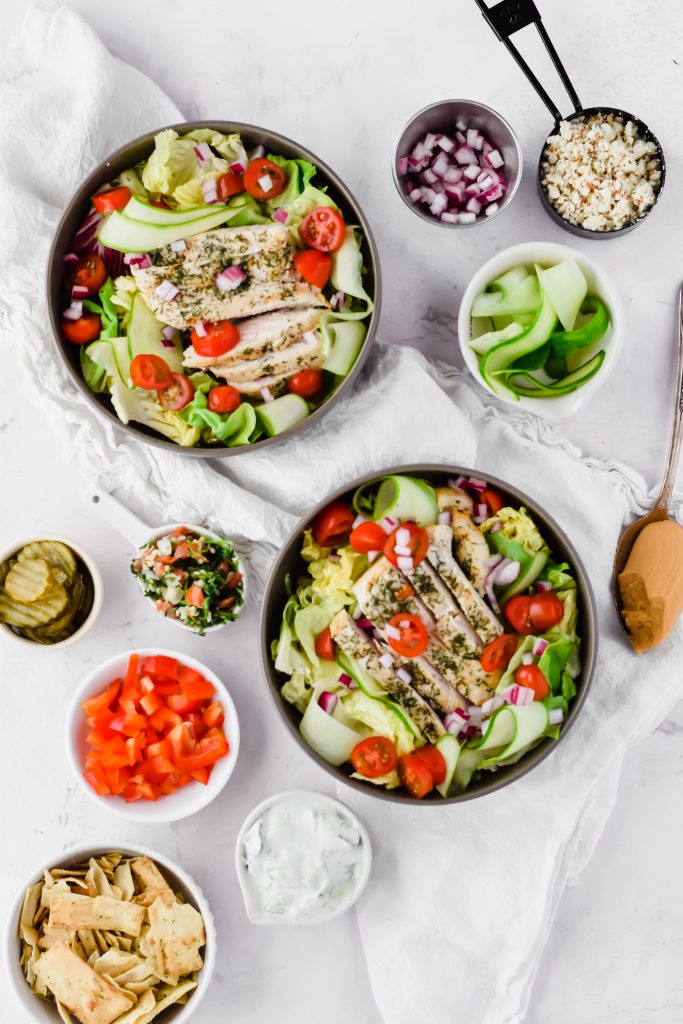 overhead shot of mediterranean chicken salad on white background beside additional bowls of chopped peppers, tabouleh, onions, cucumber, pita chips, and pickles