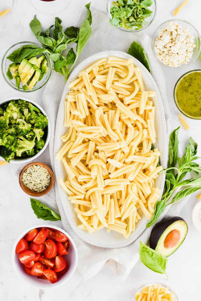 platter of cooked noodles with fresh ingredients of avocado, basil, tomato, broccoli, and feta surrounding it