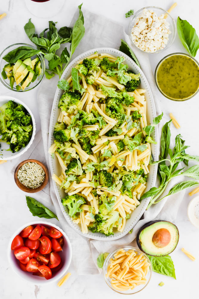 cooked pasta on oval shaped platter mixed with broccoli and chopped kale with small bowls of other ingredients surrounding it
