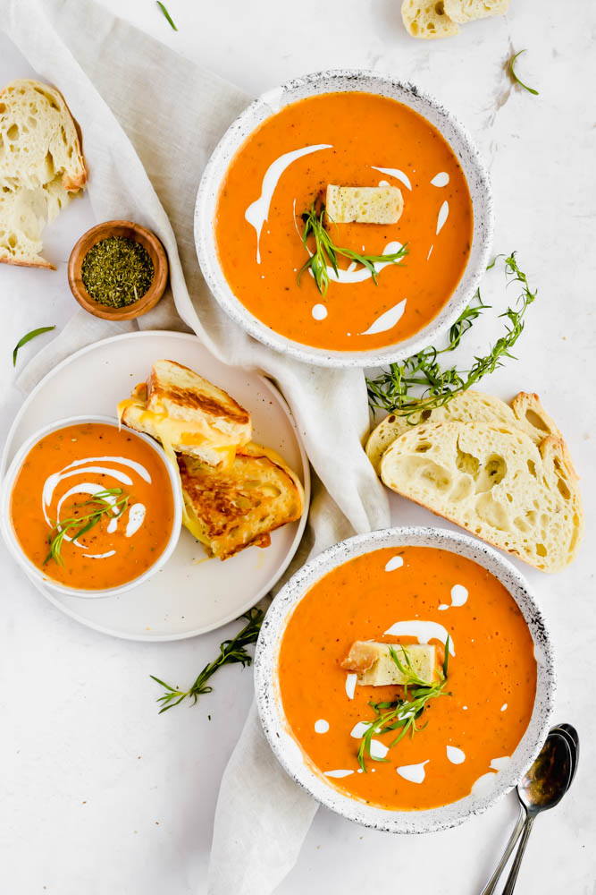 three bowls of tomato soup with grilled cheeses and slices of bread on white background