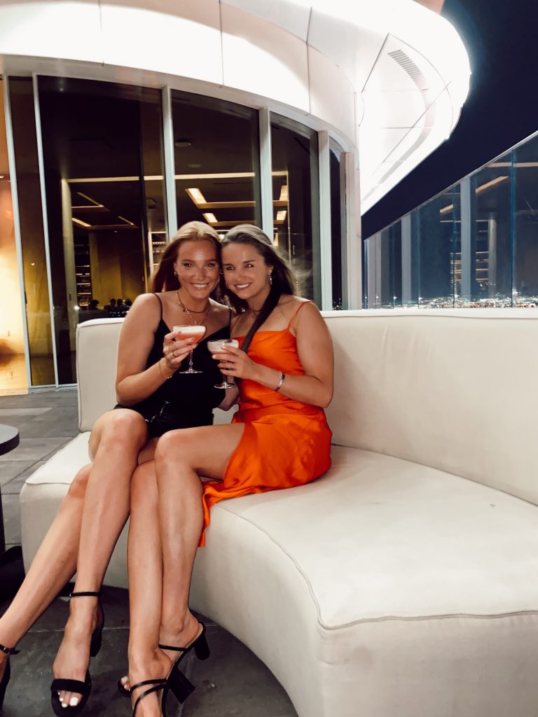 two girls in club attire sitting on white booth holding drinks overlooking skyline