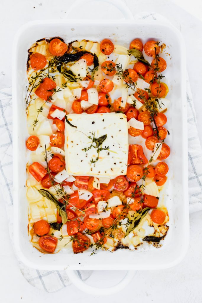 Large white baking dish filled with roasted onion, sliced cherry tomatoes, thyme sprigs, and a block of feta in the center on white and gray oven mit