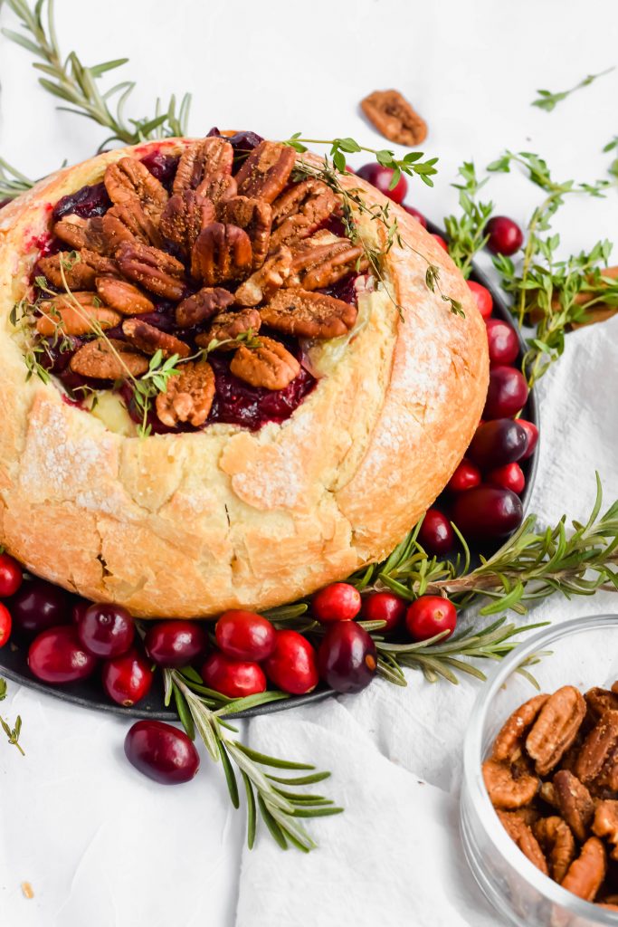 finished cranberry brie bread bowl on slate plate with fresh rosemary, cinnamon, pecans, and cranberries surrounding on white background