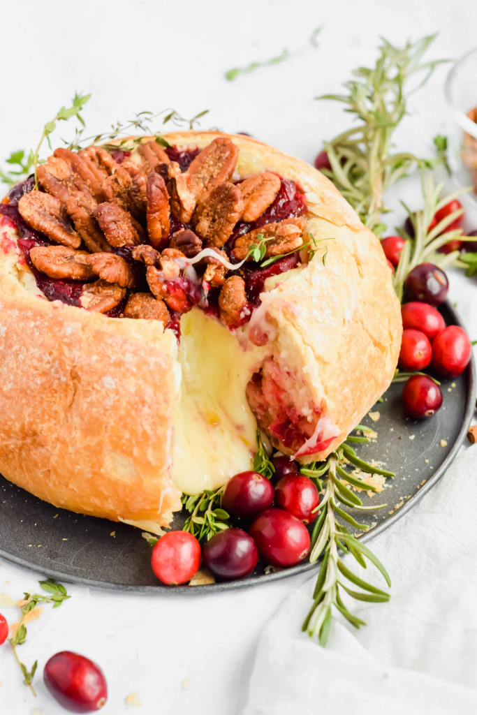 finished cranberry brie bread bowl with slice taken out on slate plate with fresh rosemary and cranberries surrounding on white background