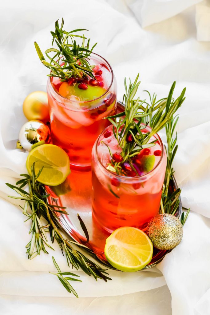 two cranberry fizz cocktails garnished and surrounded by fresh rosemary and lime slices on silver tray with white background 