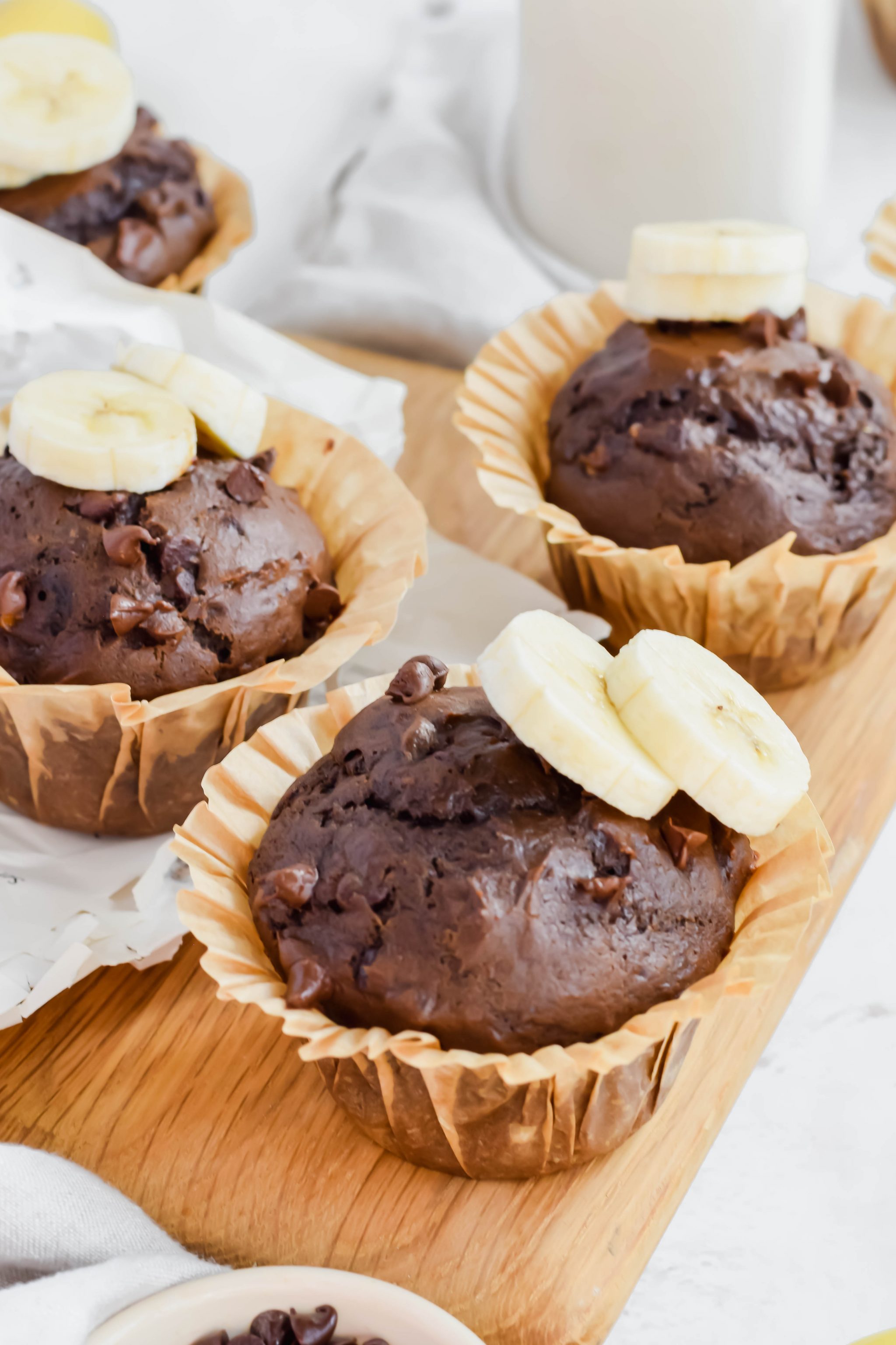 Three chocolate banana muffins topped with fresh banana slices in tan parchment liners on wood board