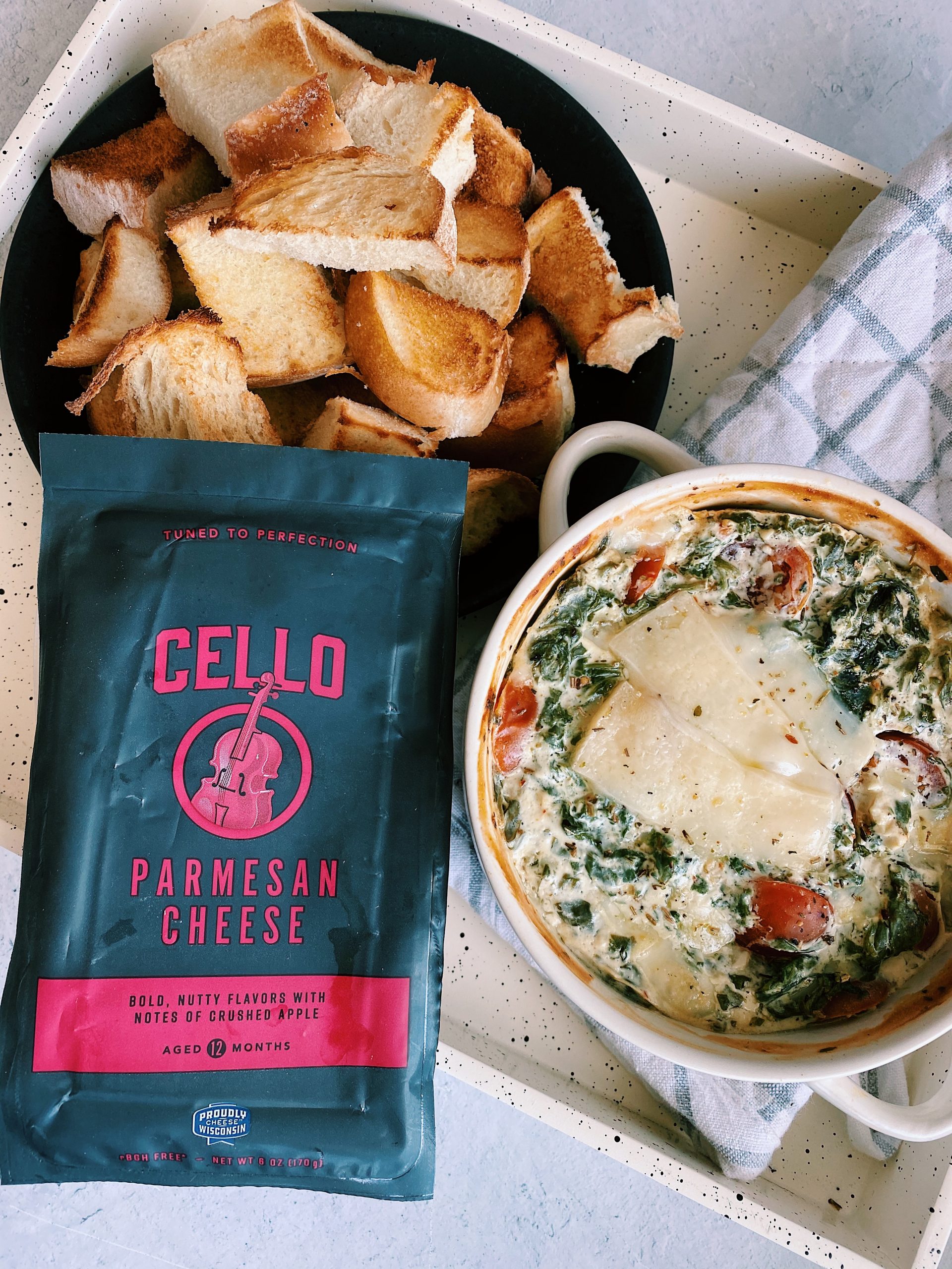 baked spinach and roasted tomato dip beside pieces of crusty bread and block of Cello parmesan cheese.