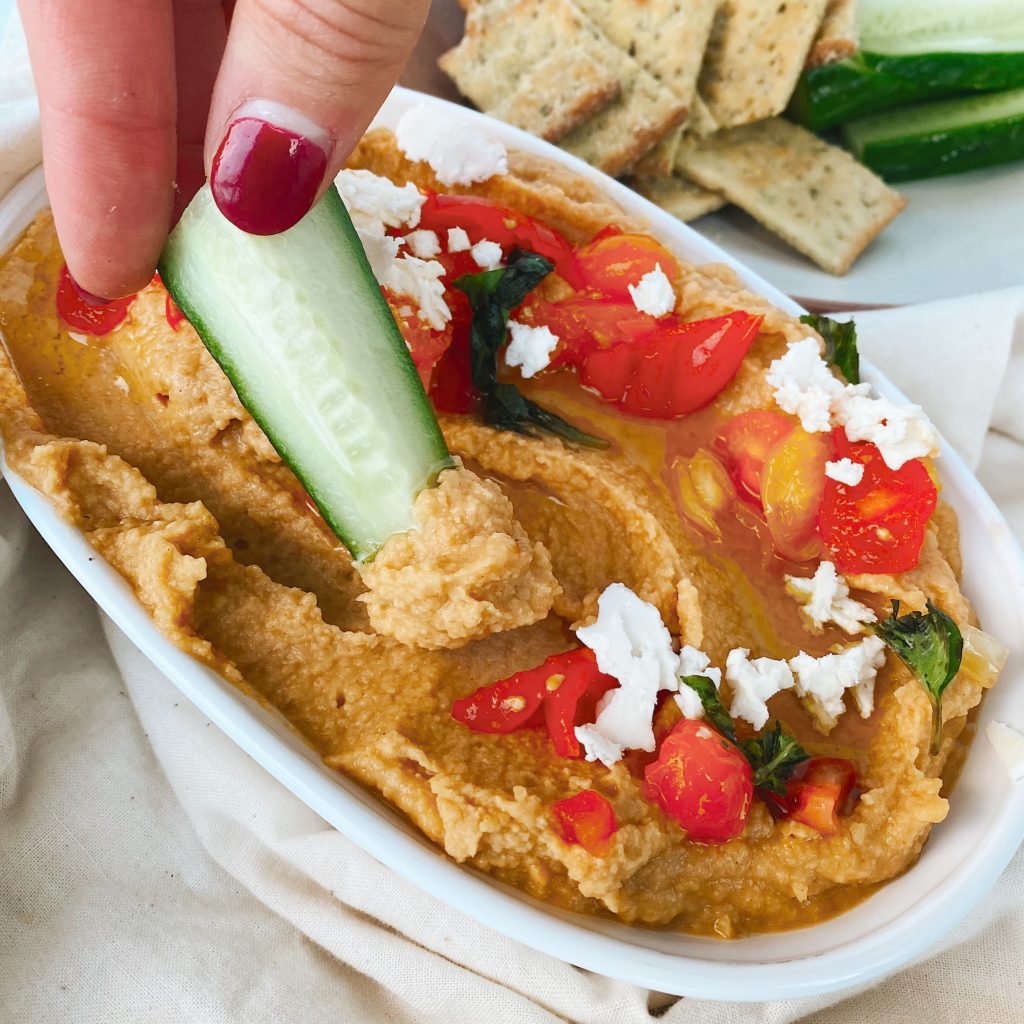 close up image with hand holding cucumber dipping through hummus