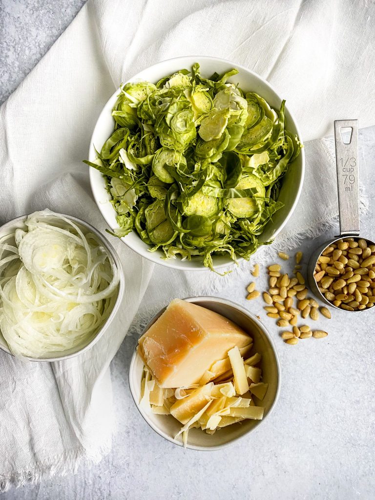 flatly shot of all ingredients laid out for roasted brussel sprout salad including brussel sprouts, pine nuts, white onions, and parmesan cheese
