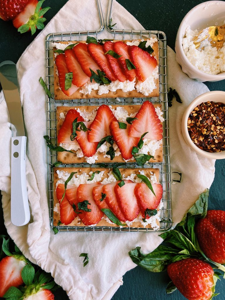 overhead flay lay shot of 3 crackers with crumbles of cheese topped with thin sliced strawberries with basil garnish all laying on top of metal wire rack