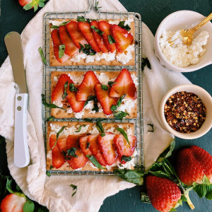 overhead flay lay shot of 3 crackers with crumbles of cheese topped with thin sliced strawberries with basil garnish all laying on top of metal wire rack