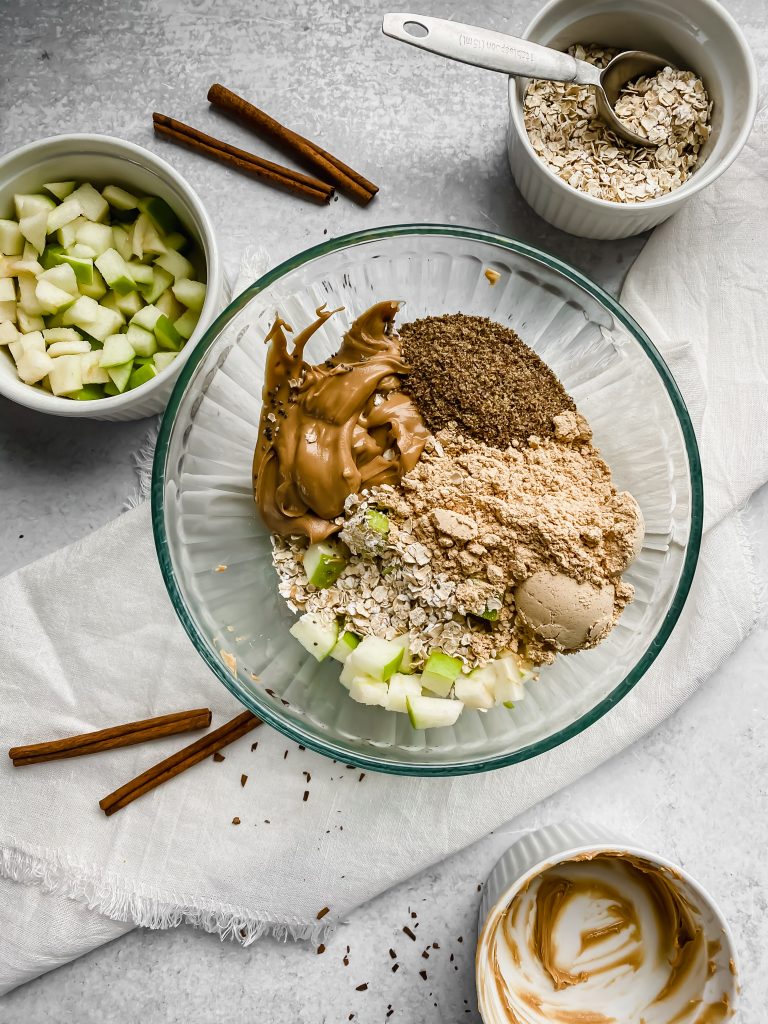 A glass bowl if filled with the ingredients for apple peanut butter energy bites. There's powdered peanut butter, oats, cinnamon, flaxseed, chia seeds, peanut butter, and diced apples. There are more bowls in the background filled with diced apples, rolled oats, and peanut butter. Cinnamon sticks are scattered around.
