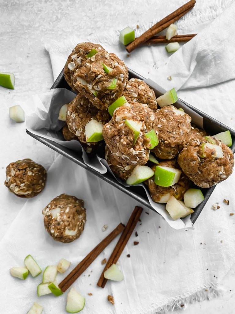 A pan is lined with parchment paper and filled with apple peanut butter energy bites. There are more energy bites, diced apples, and cinnamon sticks scattered around the pan.