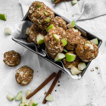 A pan is lined with parchment paper and filled with apple peanut butter energy bites. There are more energy bites, diced apples, and cinnamon sticks scattered around the pan.