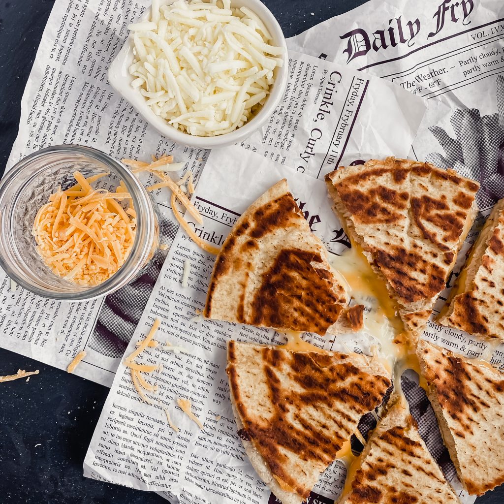 A cheesy quesadilla is sliced into six pieces with melty cheese oozing out. There is more shredded mozzarella and cheddar in the background.