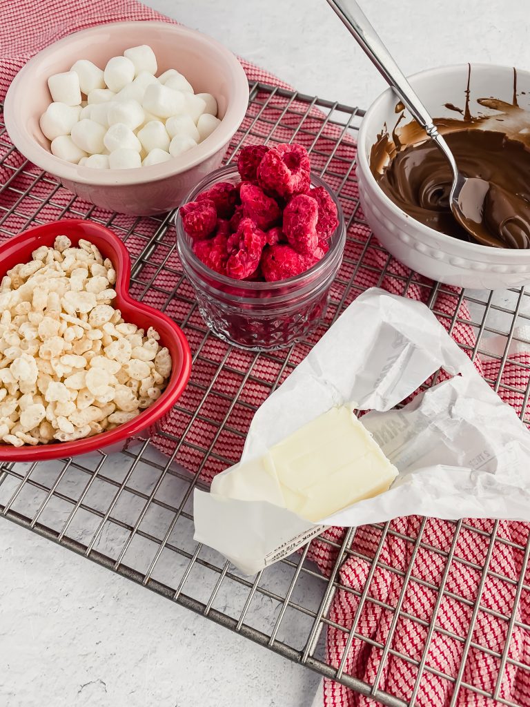 Ingredients for raspberry chocolate rice krispies including rice krispies cereal, butter, raspberries, chocolate and marshmallows, on silver rack a top a red dish rag