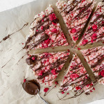 Six slices of Raspberry Chocolate Rice Krispies sit in a circle on parchment paper. A spoon covered in melted chocolate sits beside them to drizzle chocolate overtop.