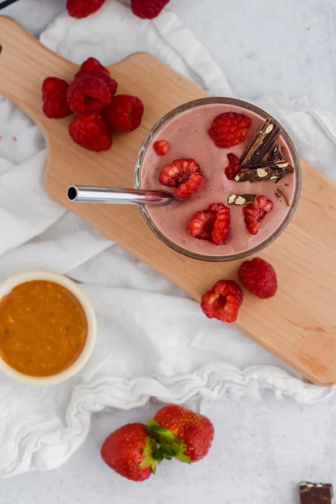 A chocolate raspberry smoothie is garnished with fresh raspberries and chocolate with a metal straw to drink it. The smoothie sits on a wooden display board with more raspberries piled around it. A white cloth sits beneath the board and there's a bowl of hazelnut butter and strawberries.