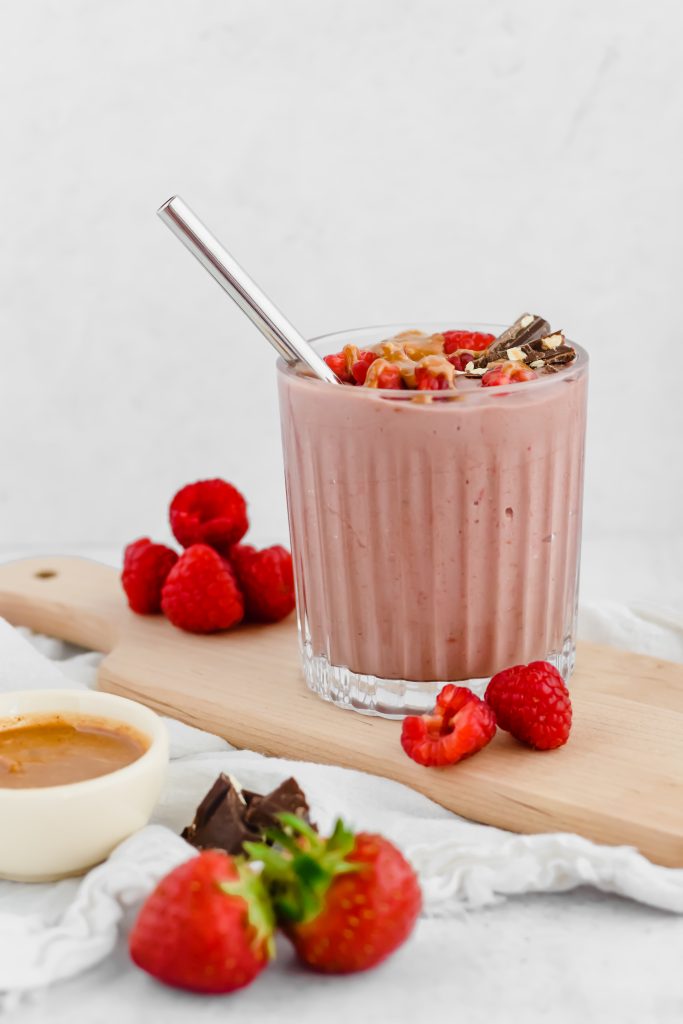 A chocolate raspberry smoothie in a glass tumbler is garnished with fresh raspberries, chocolate, and hazelnut butter drizzled overtop. The smoothie sits on a wood board with more raspberries beside it. A bowl of hazelnut butter, some chocolate pieces, and fresh strawberries sit to the side.