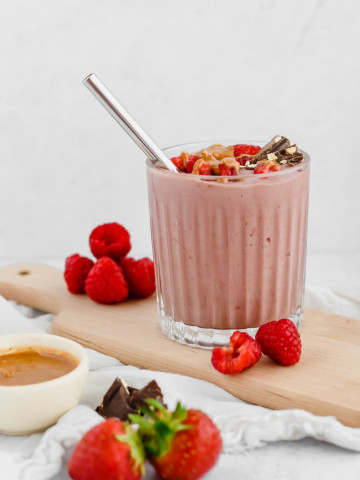 A chocolate raspberry smoothie in a glass tumbler is garnished with fresh raspberries, chocolate, and hazelnut butter drizzled overtop. The smoothie sits on a wood board with more raspberries beside it. A bowl of hazelnut butter, some chocolate pieces, and fresh strawberries sit to the side.