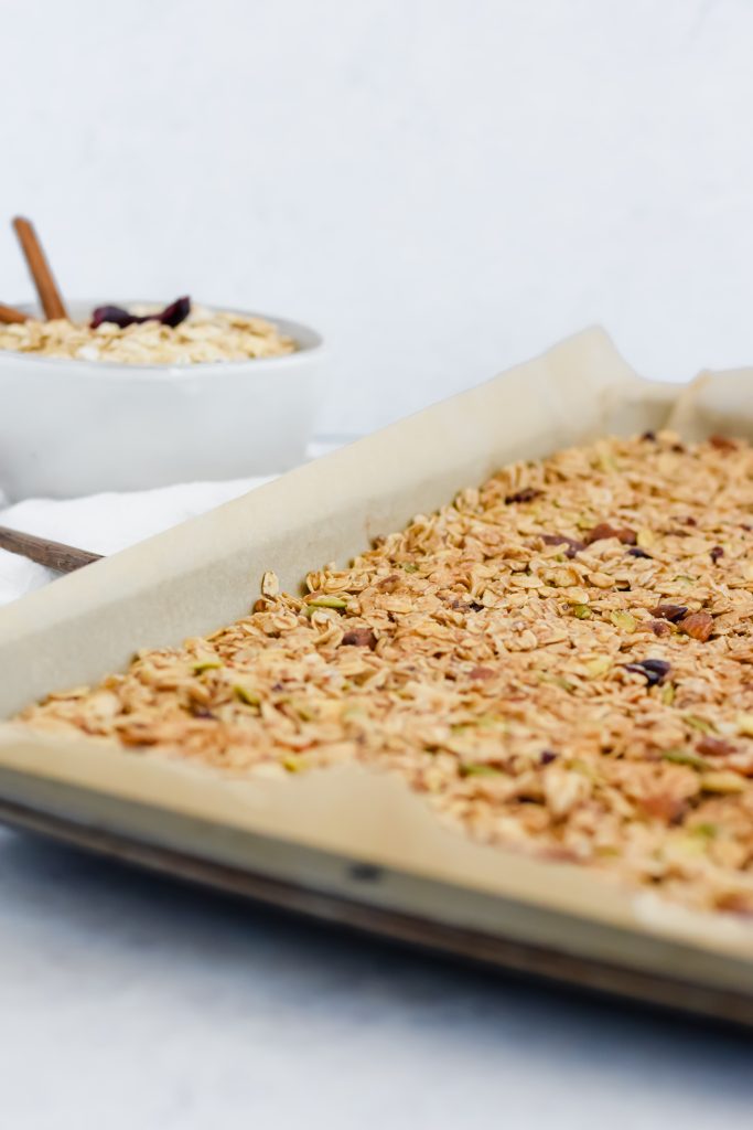 side angle shot of homemade cranberry nut granola on a parchment paper lined sheet pan on white backdrop with a bowl of granola in the background