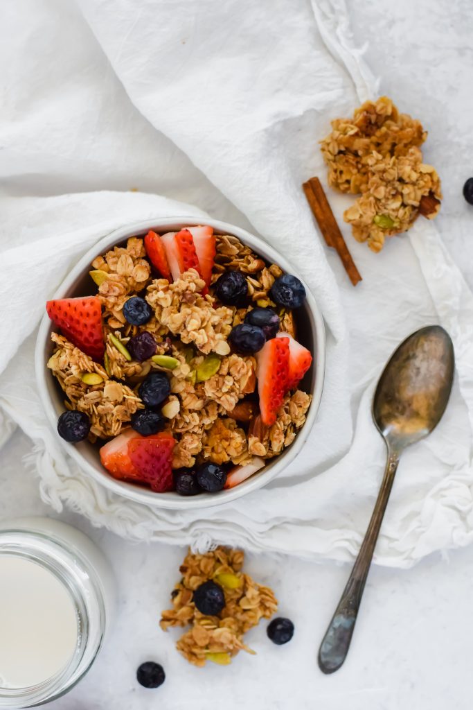 overhead shot of bowl of homemade cranberry nut granola with fresh strawberries and blueberries on white background beside a silver spoon