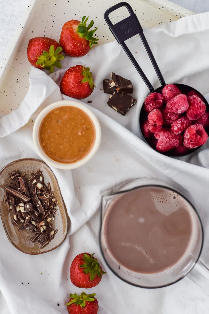 The ingredients for a chocolate raspberry smoothie all spread out on a white linen lined serving tray. There are frozen raspberries, fresh strawberries, chocolate, hazelnut butter, and a hazelnut protein shake.
