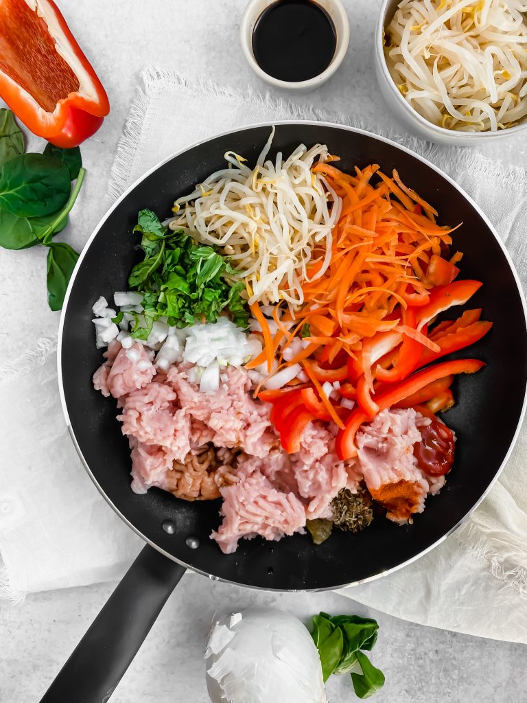 A skillet is filled with ingredients for thai chicken lettuce wraps including fresh shaved carrots, sliced red pepper, bean sprouts, ground chicken, greens, and the ingredients for a simmer sauce. There are more bean sprouts, greens, and red pepper in the background as well as a small bowl of soy sauce.