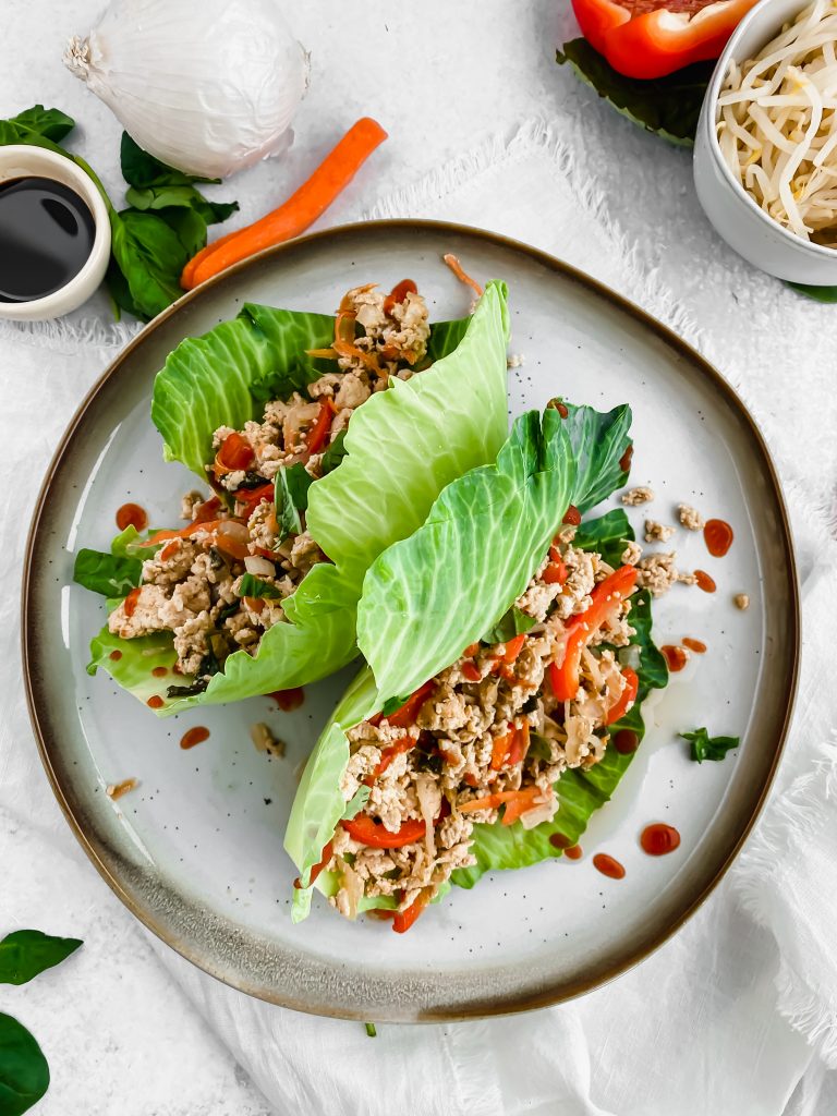Two Thai chicken lettuce wraps sit on a ceramic plate with sriracha drizzled overtop. The wraps are filled with a chicken and vegetable mixture cooked in a soy sauce simmer. There are more veggies in the background.