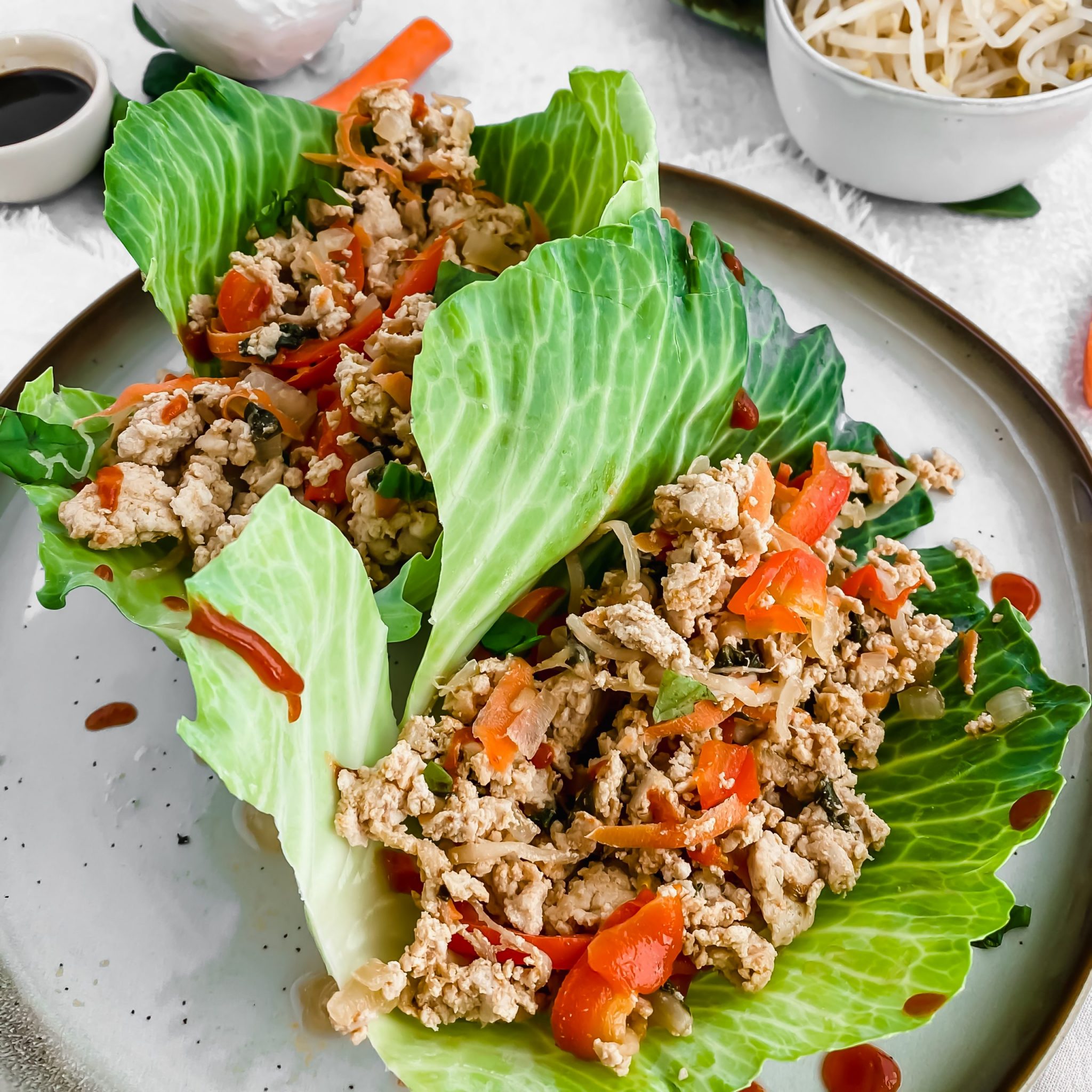 Close up of two Thai chicken lettuce wraps sitting on a ceramic plate with sriracha drizzled overtop. The wraps are filled with a chicken and vegetable mixture cooked in a soy sauce simmer. There are more veggies in the background.