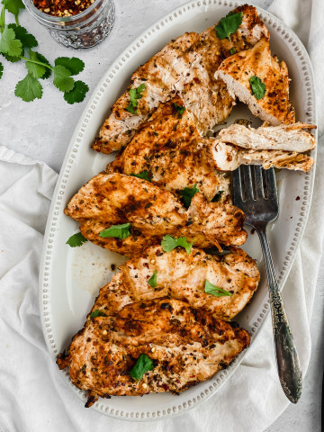 A platter is filled with the best grilled chicken breasts. A large serving fork is piercing a section of the chicken to show the juicy and tender texture. There's a white linen beneath the platter and a jar of red pepper flakes to the side of it.