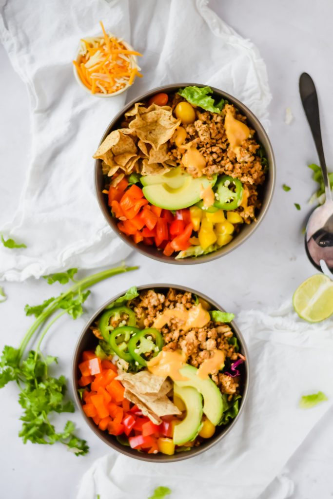 two nacho taco bowls filled with fresh veggies, cooked meat, and topped with cilantro and lime pieces on white background