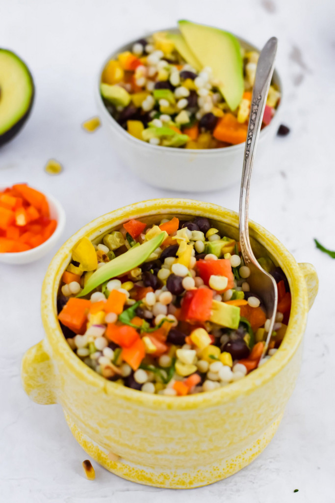 couscous salad topped with fresh avocado in a yellow ceramic bowl with spoon inside