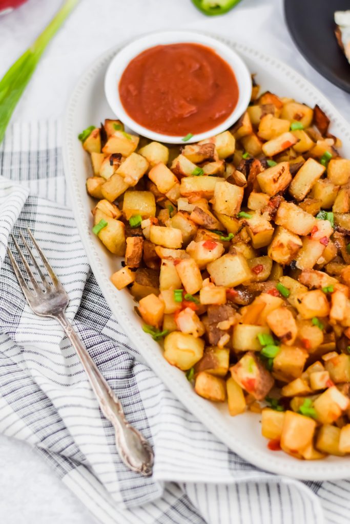 close up image of diced breakfast potatoes with scallions and salt to top and ketchup bowl on the side