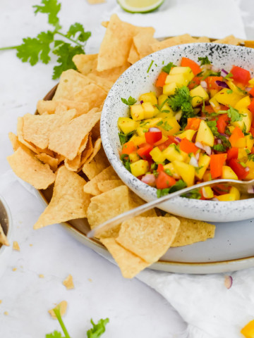 45 degree angle image of white ceramic bowl holding combined mango salsa with chips surrounding it