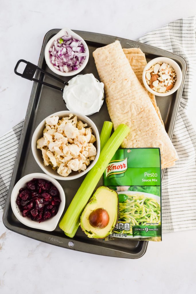 Ingredients for pesto chicken salad wrap including chicken, dried cranberries, celery, avocado, almonds, red onion, pesto seasoning, and pita on a gray sheet pan laying on top of a striped dish towel