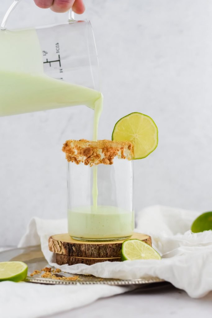 Pouring Key Lime Pie Milkshake into a glass rimmed with crumbled graham cracker and a slice of lime