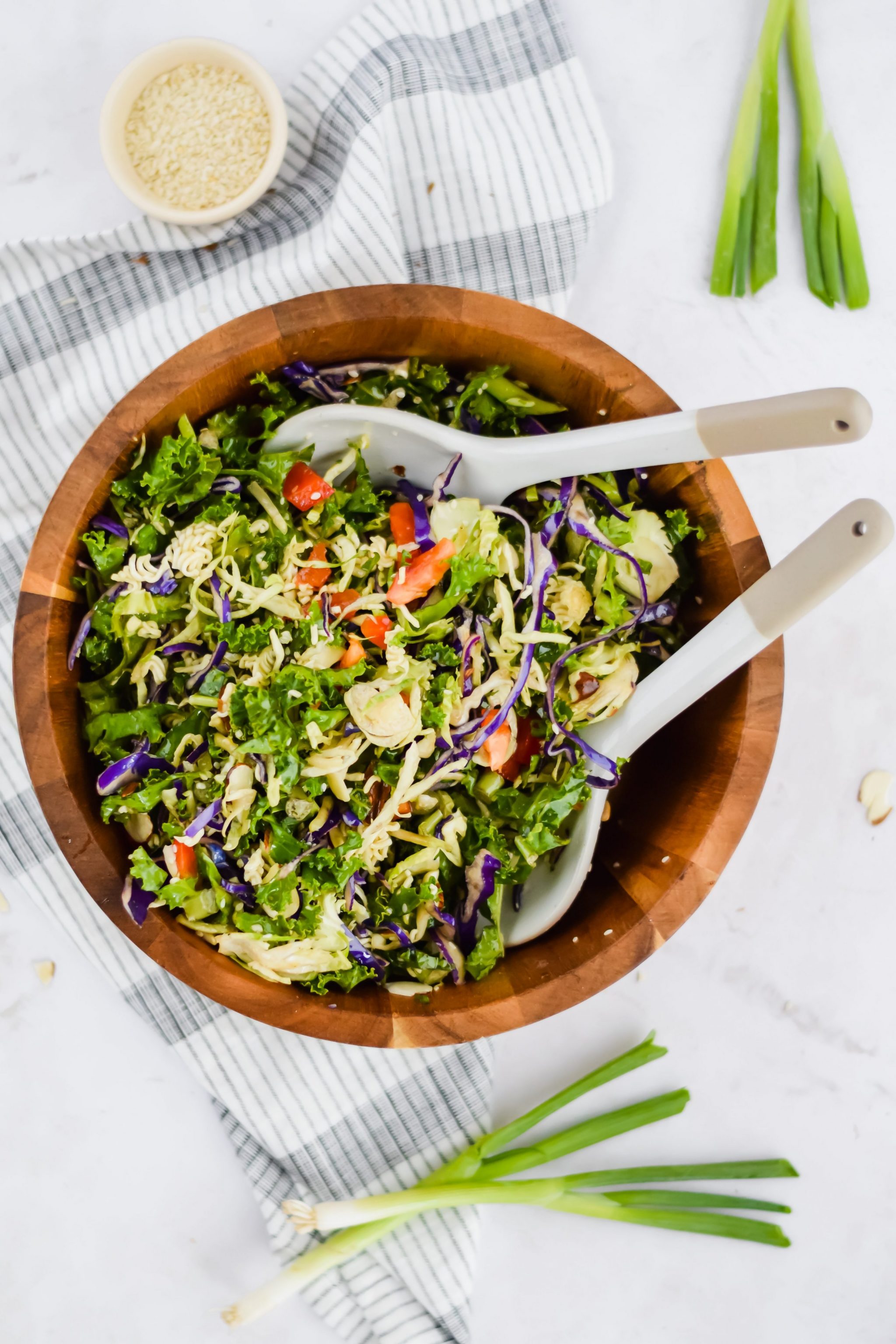 tossed oriental coleslaw salad with tongs in wooded bowl with veggies surrounding it.