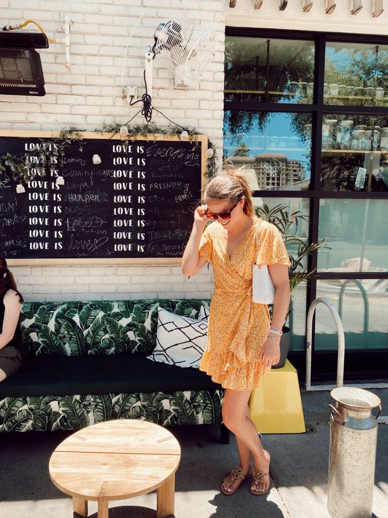 girl holding her sunglasses in yellow sundress and white purse in front of design wall