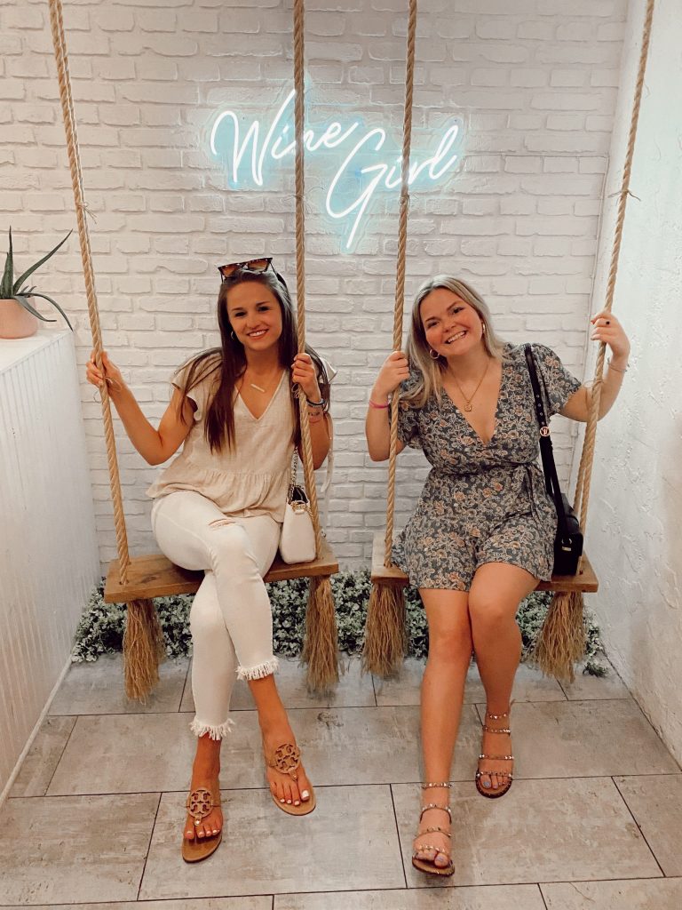 two girls in dresses on wooden swings with plants and neon sign in the background