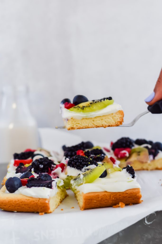 spatula holding up slice of fruit pizza with glass of milk surrounding