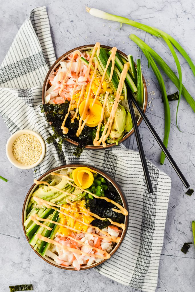 two finished california poke bowls drizzled with spicy mayo on top of striped towel and surrounded by whole green onion and small dish of sesame seeds