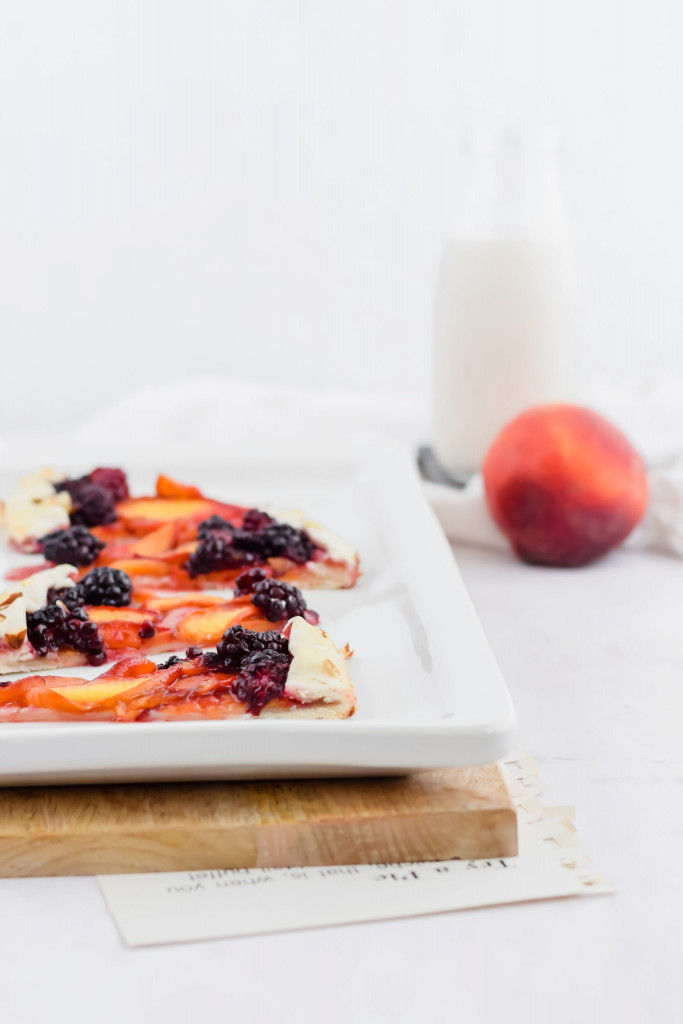 view of blackberry peach galette plated on white ceramic dish with peaches in background