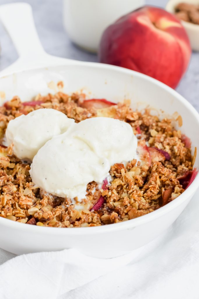close up shot of finished healthy peach crisp in white bowl topped with three scoops of vanilla icecream