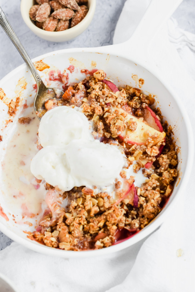 close up shot of spoon in a white bowl of healthy peach crisp with a few bites taken out topped with vanilla ice cream 